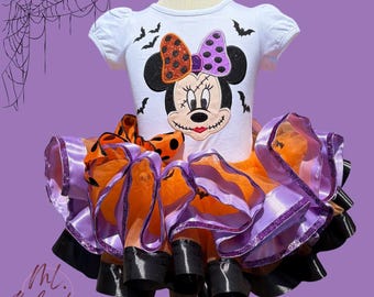 Puede incluir: Una camiseta blanca de manga corta con un gráfico brillante de Minnie Mouse con detalles cosidos y murciélagos negros, combinada con una falda de tul en capas naranja, morada y negra con ribete de satén y lunares.
