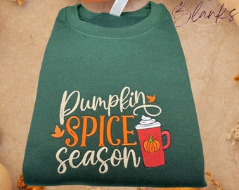 May include: A green sweatshirt with the text "Pumpkin Spice Season" embroidered in orange, white, and brown. The design also includes a small embroidered image of a pumpkin spice latte.