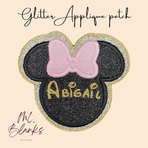 Puede incluir: Un parche de aplique de purpurina negra con forma de cabeza de ratón, con un lazo rosa y el nombre "Abigail" en letras doradas. El parche está bordeado con purpurina dorada y plateada. El texto "Glitter Applique patch" está en la parte superior.