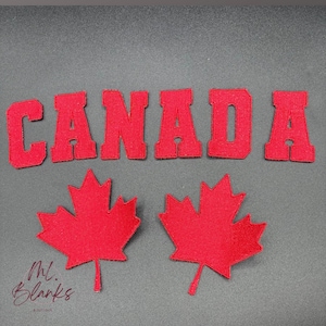 May include: Red felt letters spelling "CANADA" with two red felt maple leaf shapes. The letters and leaves are on a grey background. The bottom left corner has the text "Ml. Blanks Boutique".
