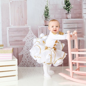 May include: A young child wearing a white dress with gold accents stands in front of a pink rocking chair. The child is holding onto the back of the chair and looking at the camera. The background is a pink and white wall with a white wooden crate and a white Christmas tree.