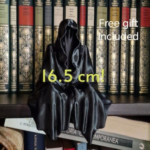Może przedstawiać: Czarna figurka postaci z kapturem wykonana z gładkiego, błyszczącego materiału. Figurka siedzi z rękami na kolanach. Figurka ma 16,5 cm wysokości i stoi na półce z książkami z tyłu. Tekst "Darmowy prezent w zestawie" jest widoczny na obrazku.