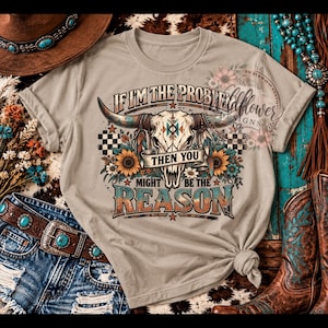 May include: A beige t-shirt featuring a graphic of a longhorn skull adorned with feathers and sunflowers. The text reads, IF I'M THE PROBLEM THEN YOU MIGHT BE THE REASON. The design includes checkered patterns, stars, and a distressed colour palette.