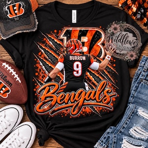 Könnte beinhalten: Ein schwarzes T-Shirt mit einer Grafik eines Footballspielers, der ein schwarzes Trikot mit dem Namen BURROW und der Nummer 9 trägt, umgeben von orangefarbenen Krallenspuren, dem Wort Bengals in orangefarbener Schrift und einem Blumenlogo mit Wildflower DESIGNS.