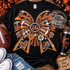 May include: A black t-shirt featuring a large graphic of a decorative bow with orange, black, and white patterns, including footballs, leopard print, and plaid. The design includes the text Bengals, Football, Who Dey, and a tiger mascot logo.