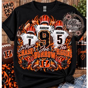 Könnte beinhalten: Ein schwarzes T-Shirt mit einer Grafik von drei Footballspielern in weißen Trikots, umgeben von orangefarbenen und schwarzen Flammen. Der Aufdruck zeigt die Namen Burrow, Chase und Higgins, die Nummern 9, 1 und 5 sowie das Bengals-Teamlogo.