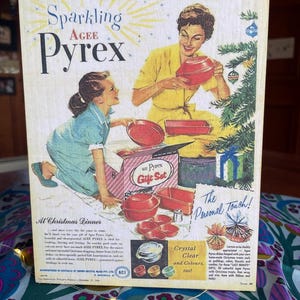 May include: A vintage advertisement for Agee Pyrex cookware featuring a woman and a girl holding red Pyrex dishes. The text reads "Sparkling Agee Pyrex", "We'll use them right away!" and "The Personal Touch!"  The ad also features a Christmas dinner setting with a Pyrex gift set.