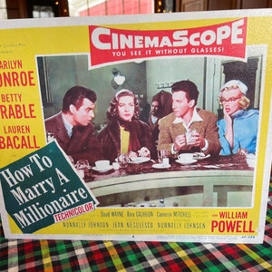 May include: A vintage movie poster for the film "How To Marry A Millionaire" starring Marilyn Monroe, Betty Grable, and Lauren Bacall. The poster is in color and features a scene from the film with the three actresses sitting at a table in a restaurant. The poster also includes the title of the film, the names of the actors, and the tagline "You See It Without Glasses!"