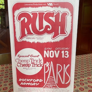 May include: A vintage concert poster advertising a Rush concert with Cheap Trick as the special guest. The concert is scheduled for Saturday, November 13th at the Rockford Armory in Rockford, Illinois. The poster is red and white with the band's name in large, stylized letters.