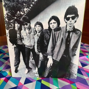 May include: Black and white photo of a band of five men standing in a row. The men are wearing casual clothing and have their hair styled in a 1970s fashion. The photo is taken in an outdoor setting.