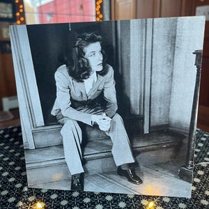 May include: A black and white photograph of a woman sitting on a staircase. She is wearing a suit jacket and pants. The woman is looking off to the side.