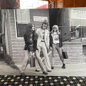 May include: Black and white photo of three people standing in front of a building. The building has a sign that says "Activities & Information Centre". The people are wearing casual clothing and are holding suitcases.