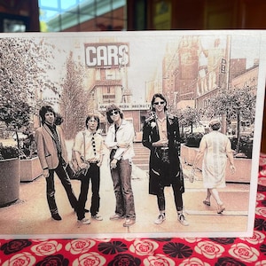 May include: A black and white photo of a band called "CARS" with five people standing in front of a building. The band members are wearing casual clothing and are looking at the camera.