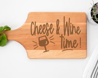 May include: A wooden cutting board with a handle, featuring the engraved text "Cheese & Wine time!" and a wine glass illustration. The board is light brown and sits on a white surface, with a plant and a striped towel in the background.