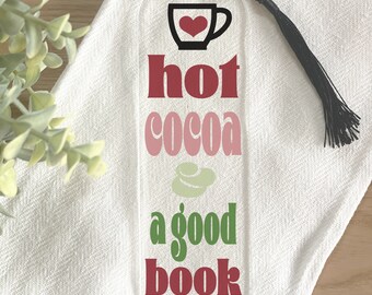 Könnte beinhalten: Transparenter Acryl-Lesezeichen mit schwarzem Quastenanhänger. Der Lesezeichen zeigt die Worte "hot cocoa a good book" in Rot, Rosa und Grün. Oben befindet sich eine schwarze Kaffeetasse mit rotem Herz.
