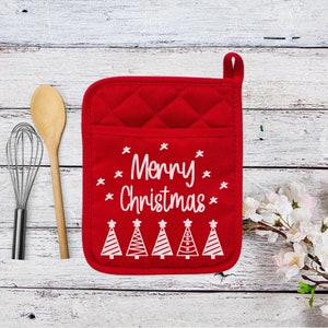 May include: Red quilted oven mitt with the text "Merry Christmas" and white Christmas tree designs. A wooden spoon and whisk are to the left of the mitt. The background is a white wooden surface.