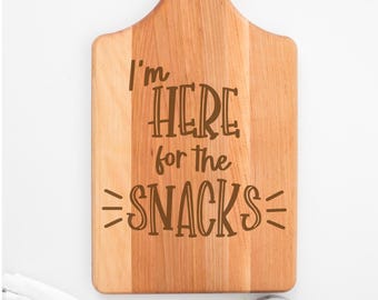 May include: Wooden cutting board with the text "I'm HERE for the SNACKS" in brown lettering. The board has a handle and is decorated with fresh basil. A striped towel and a small potted plant are in the background.