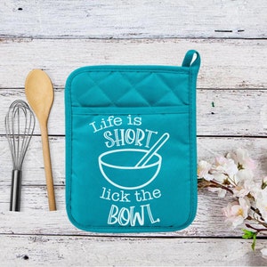 May include: Teal quilted oven mitt with the text "Life is Short lick the BOWL" in white. A wooden spoon and a whisk are to the left of the mitt. The mitt has a pocket and a loop for hanging.