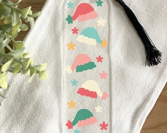 Könnte beinhalten: Transparenter Acryl-Lesezeichen mit bunten Weihnachtsmützen und Schneeflocken. Die Mützen sind in Rosa, Grün und Blau mit weißem Rand gehalten. Eine schwarze Quaste hängt oben. Ein festliches Accessoire für Leser.
