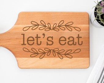 May include: A rectangular wooden cutting board with a handle, featuring the phrase "let's eat" in brown lettering, framed by decorative leaf designs. The board is light brown with a natural wood grain.