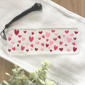 May include: A clear acrylic bookmark with a pink and red heart pattern. The bookmark has a black tassel.