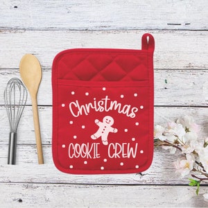 May include: A red quilted oven mitt with the words "Christmas COOKIE CREW" and a gingerbread man graphic. A wooden spoon and whisk are to the left of the mitt. White polka dots are scattered around the text.