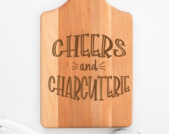 May include: A wooden cutting board with a handle, featuring the words "CHEERS and CHARCUTERIE" in a brown, decorative font. The board is light brown with a natural wood grain. A white and gray striped cloth and a small potted plant are in the background.