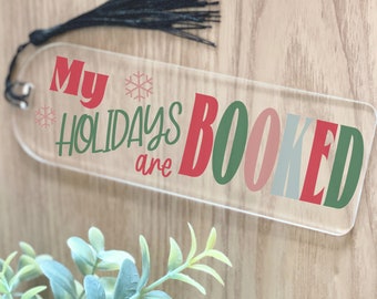 Könnte beinhalten: Transparenter Acryl-Lesezeichen mit dem Text "My Holidays are BOOKED" in Rot, Grün und Rosa. Das Lesezeichen hat eine schwarze Quaste und Schneeflocken-Akzente. Ein festliches Accessoire für Bücherliebhaber.