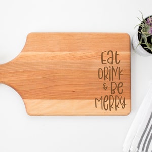 May include: A wooden cutting board with a handle, featuring the text "Eat Drink & Be Merry" in brown lettering.