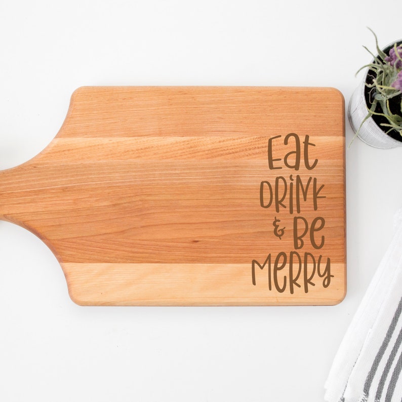 May include: A wooden cutting board with a handle, featuring the text "Eat Drink & Be Merry" in brown lettering.
