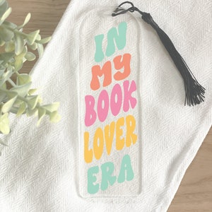 May include: A clear acrylic bookmark with a black tassel. The bookmark has the text "In My Book Lover Era" in colorful, wavy letters.