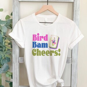 May include: White t-shirt with the words "Bird Bam Cheers!" in pink, blue, and green. A small graphic of a bird and flowers is next to the word "Cheers!". The shirt is hanging on a wooden hanger.