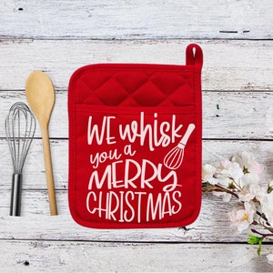 May include: Red quilted oven mitt with the text "WE whisk you a MERRY CHRISTMAS" in white. A wooden spoon and a whisk are next to the mitt. The mitt has a pocket and a loop for hanging.