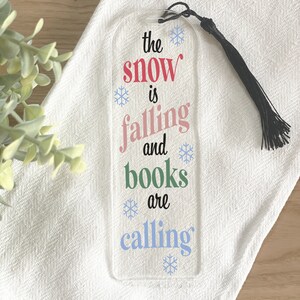 May include: Clear acrylic bookmark with the text "the snow is falling and books are calling" in red, pink, green, and blue. Includes a black tassel and snowflake accents. A decorative accessory for book lovers.
