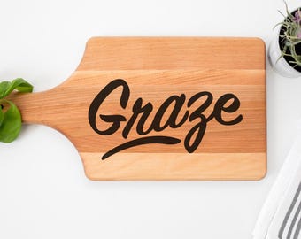 May include: A wooden cutting board with a handle, featuring the word "Graze" in a black script font. The board is light brown with a natural wood grain. A small plant and a striped towel are in the background.