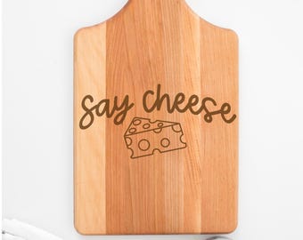 May include: A wooden cutting board with a handle, featuring the phrase "say cheese" and a cheese graphic. The board is light brown with a natural wood grain. Fresh basil and a small potted plant are in the background.