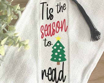 Könnte beinhalten: Transparenter Acryl-Lesezeichen mit dem Text "Tis the season to read" in Schwarz und Rot, mit einem grünen Weihnachtsbaum mit gelbem Stern. Eine schwarze Quaste hängt oben.