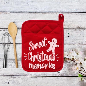 May include: A red quilted oven mitt with a pocket and a hanging loop. The mitt features the text "Sweet Christmas memories" and a gingerbread man graphic in white. A wooden spoon and whisk are to the left.