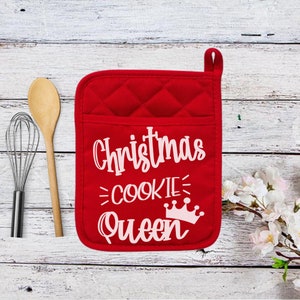 May include: Red quilted oven mitt with the text "Christmas Cookie Queen" in white. The mitt has a pocket and a loop for hanging. A wooden spoon and whisk are next to the mitt.