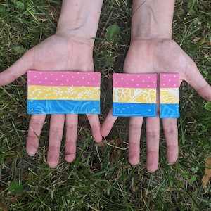 May include: Two handmade pansexual pride flags. The larger flag is pink, yellow, and blue with a white floral pattern on the yellow stripe. The smaller flag is the same design but is smaller in size.