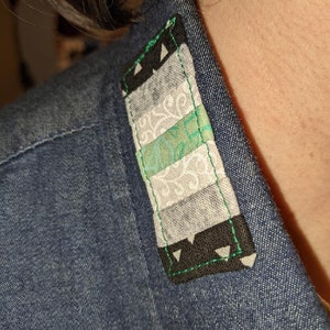 Puede incluir: Primer plano de un cuello de camisa vaquera con un peque&ntilde;o parche rectangular cosido. El parche est&aacute; hecho de tela con un patr&oacute;n geom&eacute;trico en tonos grises, verdes y negros. El parche est&aacute; bordeado con hilo verde y tiene un tri&aacute;ngulo negro en cada esquina.