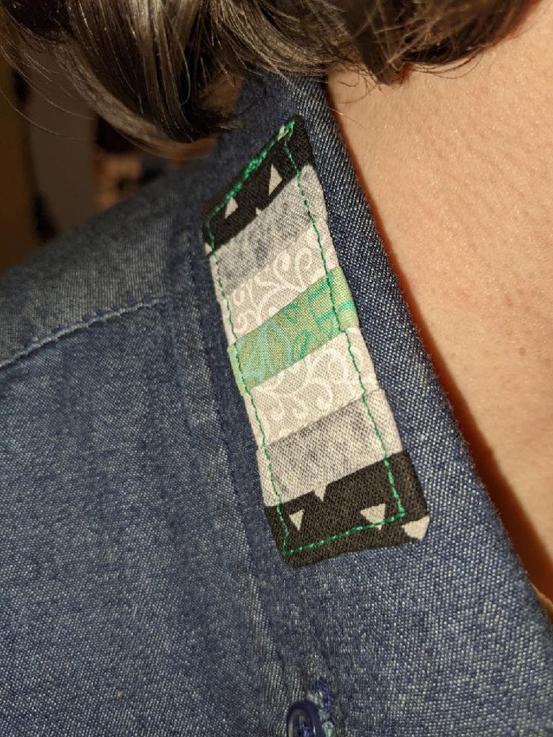 Puede incluir: Primer plano de un cuello de camisa vaquera con un peque&ntilde;o parche rectangular cosido. El parche est&aacute; hecho de tela con un patr&oacute;n geom&eacute;trico en tonos grises, verdes y negros. El parche est&aacute; bordeado con hilo verde y tiene un tri&aacute;ngulo negro en cada esquina.