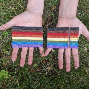 Op de afbeelding: Twee stoffen kaarten ter grootte van een portemonnee met een regenboog-pride-vlagontwerp. De kaarten zijn gemaakt van verschillende stoffen en hebben een patchwork-look. De kaarten worden in de handen van een persoon gehouden.