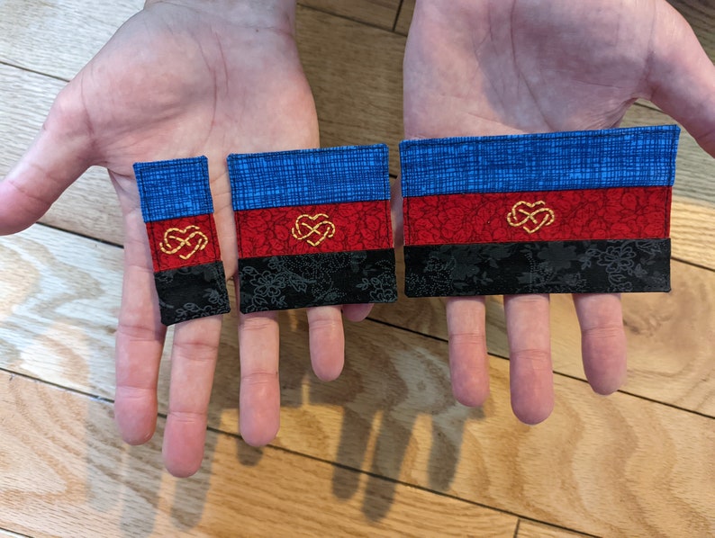 Puede incluir: Tres parches de tela de diferentes tama&ntilde;os, cada uno con un esquema de color azul, rojo y negro con un s&iacute;mbolo de infinito dorado en el centro. Los parches est&aacute;n hechos de una tela texturizada y tienen un brillo sutil.