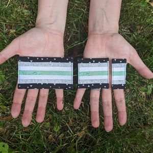 Puede incluir: Tres parches de tela de diferentes tamaños, cada uno con un fondo negro y triángulos blancos. Los parches tienen un patrón de rayas verdes y blancas, que se asemeja a una bandera del orgullo.