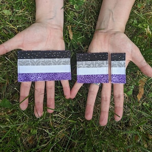 May include: Two fabric patches featuring the asexual pride flag design. The larger patch is approximately 7.5 cm wide and the smaller patch is approximately 5 cm wide. Both patches are in a black, grey, white and purple colour scheme.
