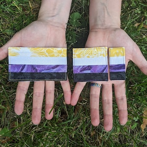 May include: Two non-binary pride flag patches, one larger than the other, are held in the palms of a person's hands. The patches are made of fabric and feature a yellow, white, purple, and black color scheme. The patches are on a green grassy background.