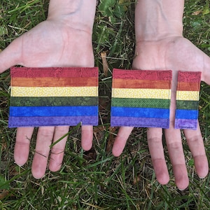 May include: Two rainbow pride flags made of fabric with a green, yellow, orange, red, blue, and purple color scheme. The flags are different sizes and are held in the hands of a person.