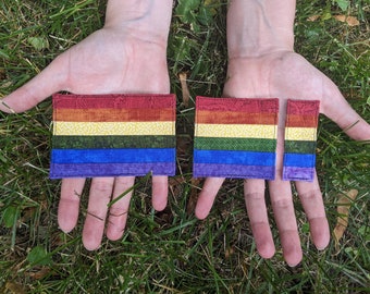 May include: Two rainbow pride flags made of fabric with a green, yellow, orange, red, blue, and purple color scheme. The flags are different sizes and are held in the hands of a person.