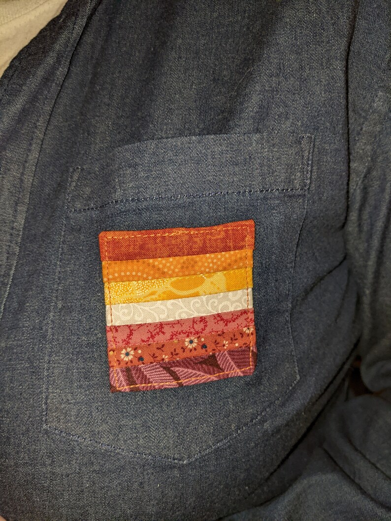 Puede incluir: Un peque&ntilde;o parche colorido cosido en una camisa vaquera. El parche est&aacute; hecho de diferentes telas y presenta un patr&oacute;n de rayas y dise&ntilde;os florales en tonos rojos, naranjas, amarillos, blancos y rosas.